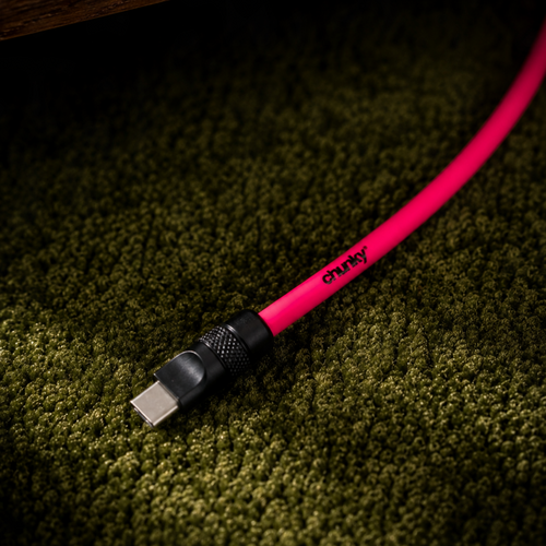 A close-up of a bright pink USB-C cable with a black connector, lying on a dark green textured carpet under a wooden surface. The brand name 'chunky' is printed in black on the cable.