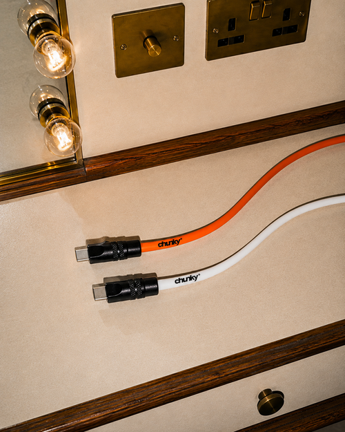 Two chunky USB-C charging cables, one orange and one white, with black connectors, laid out on a beige wall with wooden trim and light fixtures in the background.
