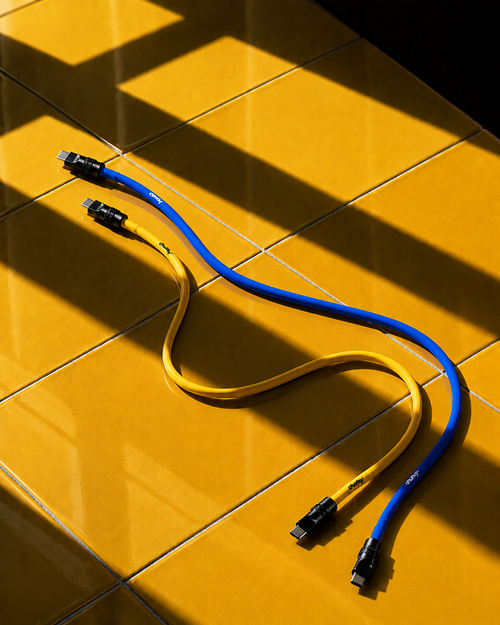 Two USB-C charging cables, one yellow and one blue, arranged in a curved loop on a glossy yellow tiled surface with dramatic diagonal shadows.